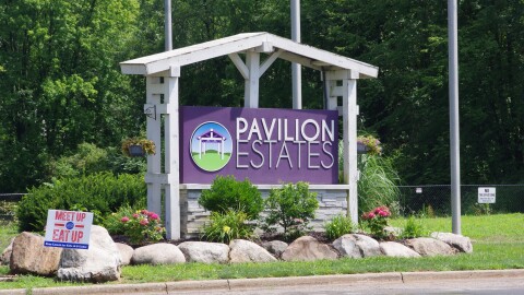 A large white and purple sign reads "Pavilion Estate." The sign is surrounded by flowers, bushes and rocks. A road passes just in front of it, with a small sign on a fence in the background reading "no trespassing."