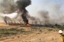 A small white and red plane flies low over a wildfire consuming brush and dry grass in a field. BLM trucks and firefighters are also around the perimeter of the fire. On firefighter is in the lower righthand corner of the image with a yellow shirt and white hardhat. 