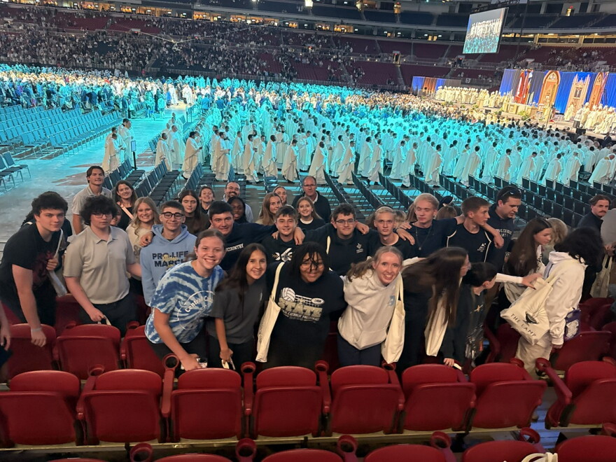 Several dozen high school students pose for a photo in an indoor arena as a gathering of men in white robes walk in a procession at a Catholic church gathering.