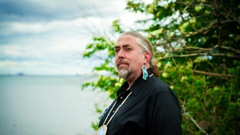 A man with a goatee, wearing a black shirt, necklace and earring stands near a body of water and green plantlife