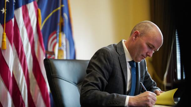 Spencer Cox signs a bill at a desk with federal and state flags behind him.