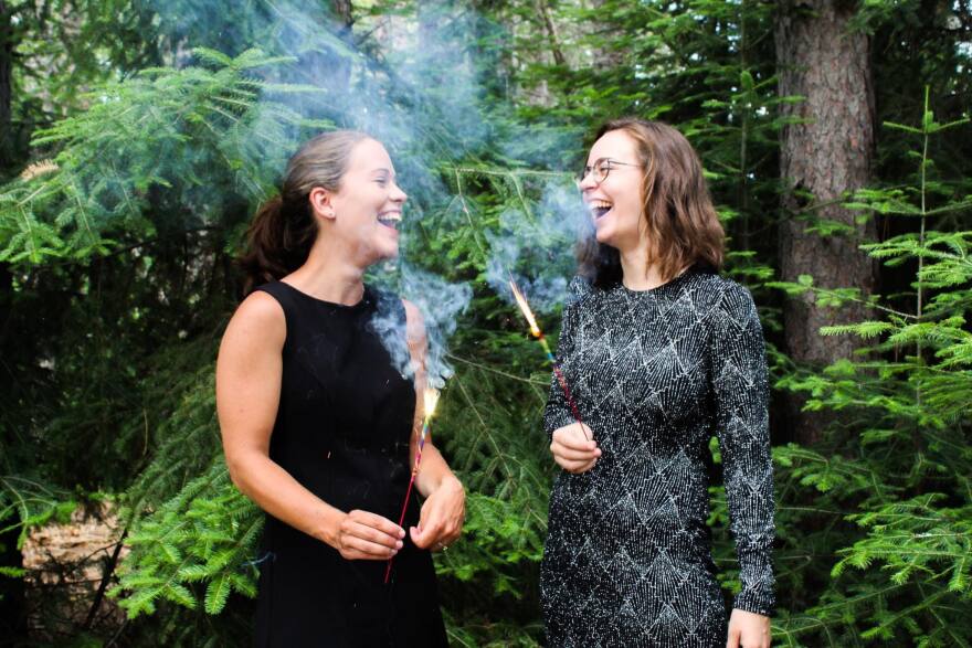 Two women laughing and looking at each. They are holding sparklers and standing in front of green trees.