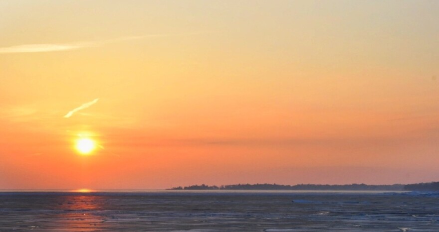 A vivid sunset over a lake with an orange, slightly clouded sky. The sun reflects on the lake. Across the lake, a forest is visible on a far-off shore. 