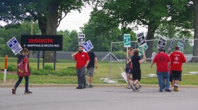 Around ten people stand on the side of the road in front of a chainlink fence and a black sign to the middle right of the image that reads "DAUCH" in red text, below which reads "Shipping & Receiving" in white text with an arrow pointing to the right. The men and women hold different colored signs, but most of them read the same message; "UAW ON STRIKE." One woman's sign to the left of the image reads "UAW ON STRIKE, UNFAIR LABOR PRACTICE" in blue text.