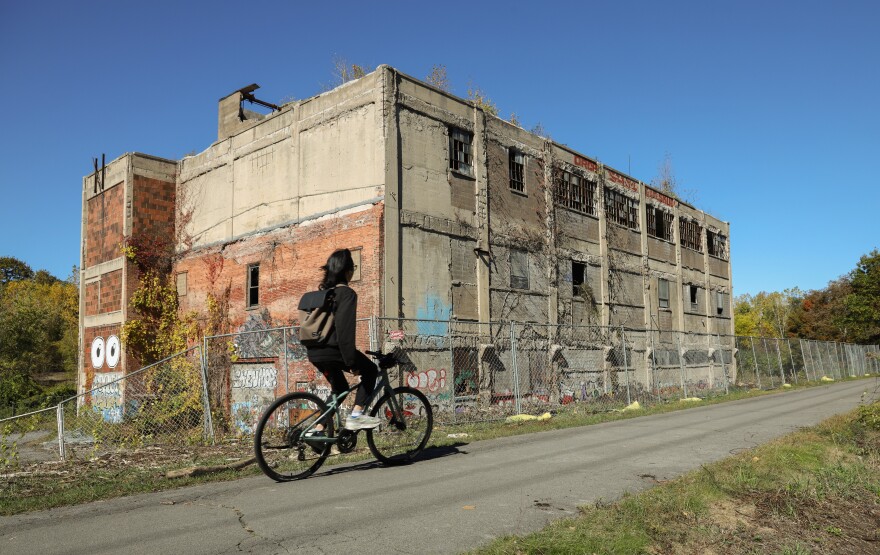 An abandoned, crumbling warehouse is in the background while a person wearing all black bicycles by on a paved trail