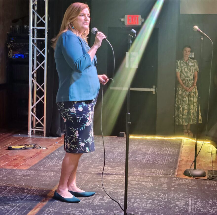 Woman holds microphone speaking on stage as another woman in long green printed dress leans on wall in corner staring at the woman on the stage 