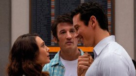 A still from the film Eternity. Elizabeth Olsen’s Joan and Callum Turner’s Luke stand close, hands clasped while Miles Teller’s Larry looks on. 