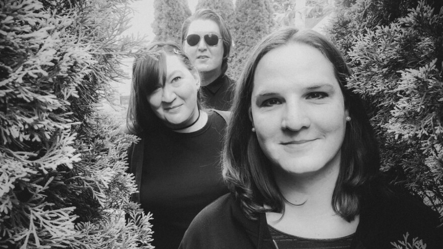 A black-and-white image of three people smiling pleasantly at the camera with fir trees on either side of them.