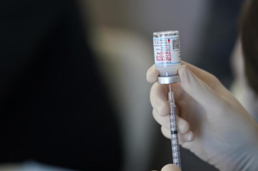 A person's gloved hand is shown inserting a syringe into a vial containing the Moderna COVID-19 vaccine.