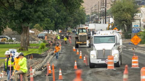 A large white truck surrounded by orange traffic cones and men in safety vests and hard hats in the foreground show construction underway on Snake River Avenue in Lewiston. 