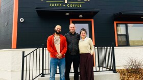 three people stand together and smile in front of a dark gray storefront that says Kuumba Juice + coffee