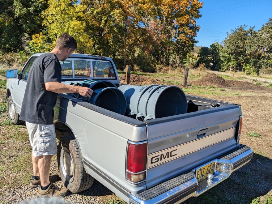 A man wearing a black shirt and white cargo shorts arranges a stack of grey bins int he back of a silver GMC pickup truck outside.