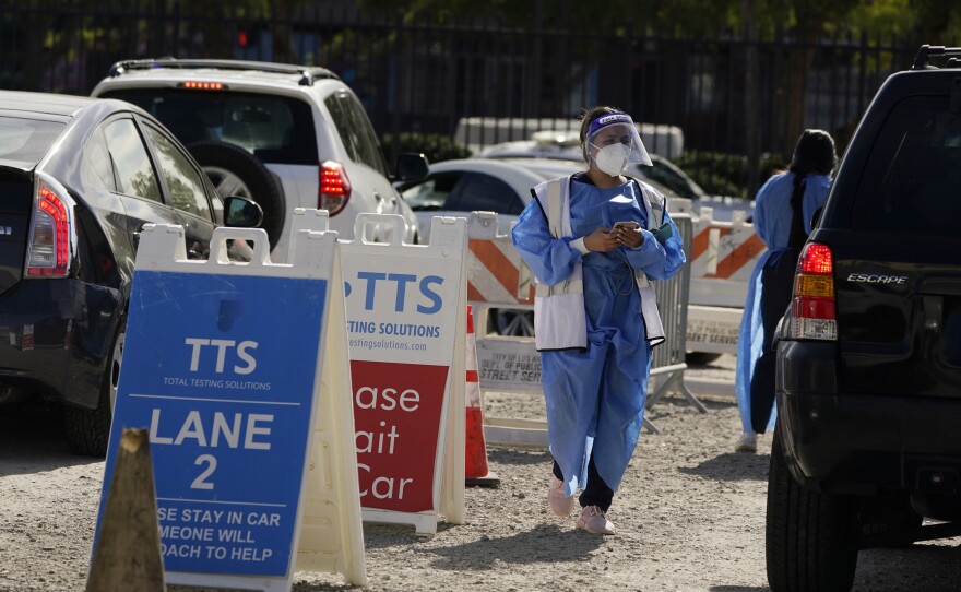 A healthcare worker wearing a face mask, face shield and blue gown walks between lanes of cars at a COVID-19 testing site. 