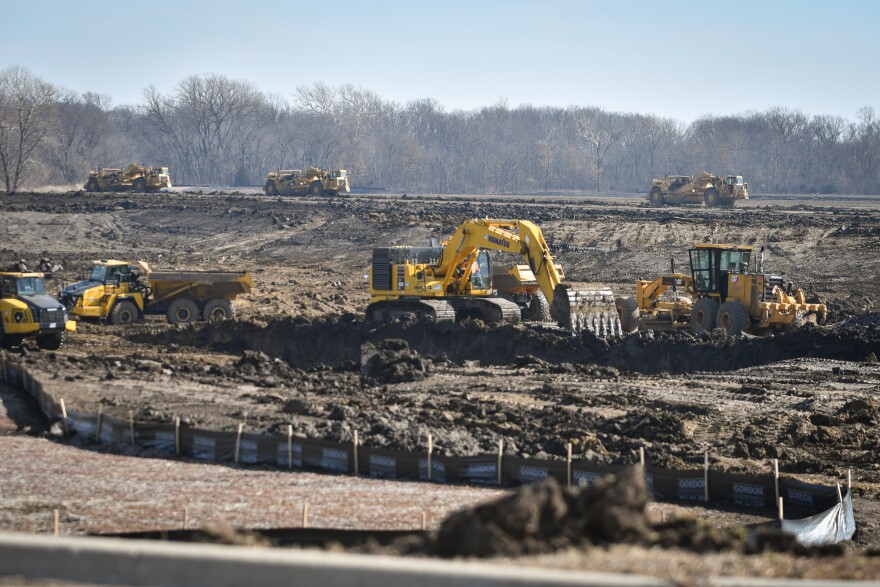 Exterior daytime image of an expansive dirt patch shows several yellow earth-moving vehicles at work.