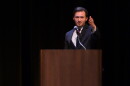 A young man in a suit extends his arm out to the crowd under a spotlight with a black background 