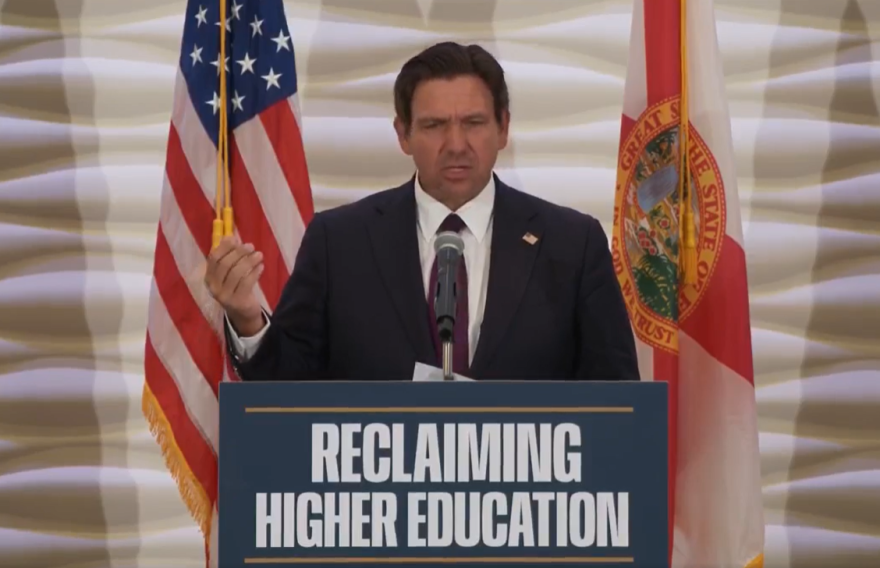 Man in a suit and tie stands behind a podium with the sign "reclaiming higher education." An American and Florida flags are behind him.