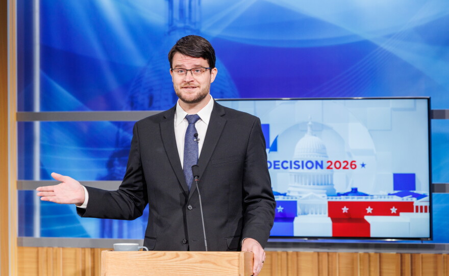 A man in a suit speaks at a podium in front of a TV that reads "Decision 2026"