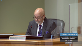 A man in a suit sits at a podium looking down with his glasses on his nose