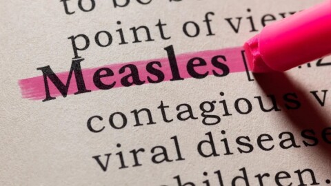 illustration represents close up of a book page with the words Measles, contagious and viral disease, with a red highlighter marking through the word Measles. 