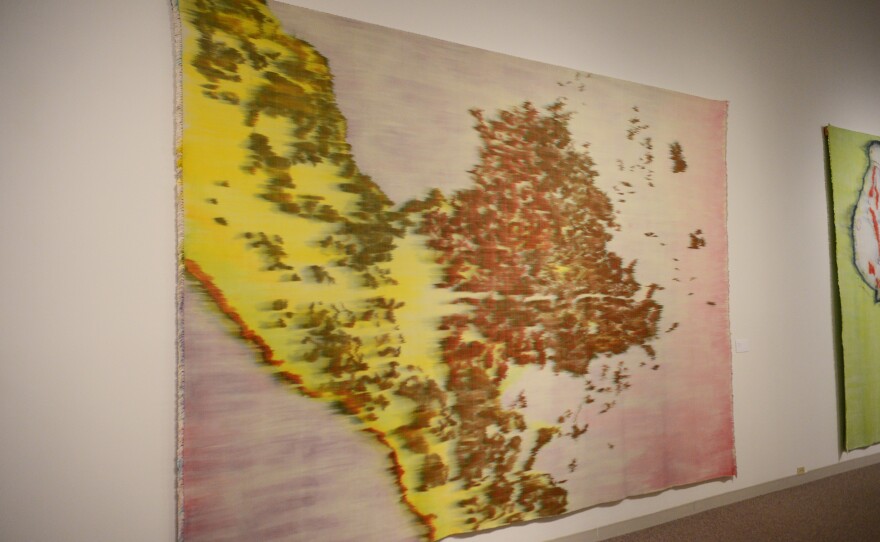 A large, colorful textile artwork hangs on the wall in a gallery