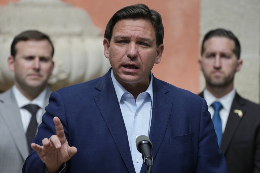 Man speaks at a microphone, two other men stand behind him.