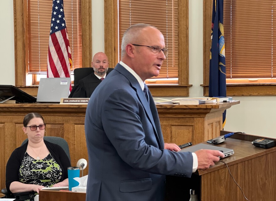 Michigan Assistant Attorney General William Rollstin stands at a lectern inside a courtroom while Judge Charles Hamlyn and a court reporter look on.