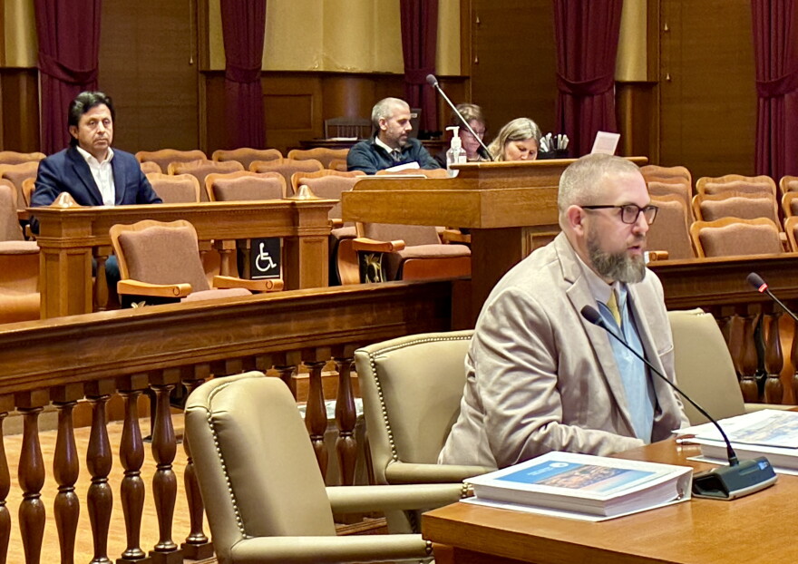 A man with a beard and glasses sits at a table in a courtroom, with other people seated in the background.