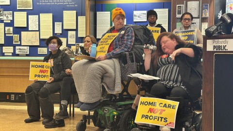 Janine Hunt-Jackson (middle, yellow hat) watches as Caring Majority activist Renee Christian speaks into a microphone at a rally at Buffalo and Erie County's Central Library. Both are using power wheelchairs. They are surrounded by other activists holding signs.