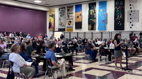 About 150 people in chairs listen to a woman standing at a podium inside an elementary school gymnasium, with musical posters on the wall.
