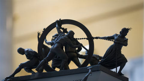 A close up of a mini model of people on top of a platform that reads "In God We Trust." The figures on top of the platform are pulling and pushing a hoop.