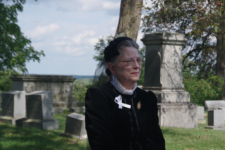 Woman in black period clothes with white collar and cap looks out of frame with grave markers in the background, and trees