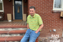 Michael Hickey sits outside his home. He wears a green striped shirt and jeans in front of a brick house.
