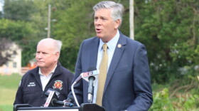 Darin LaHood speaks at the Bloomington Parks & Recreation podium as Mayor Dan Brady stands behind him in his fire department jacket.