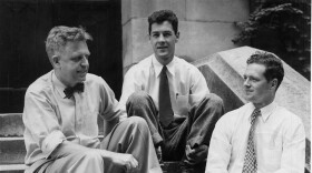 A historic black-and-white photograph of sexologist Alfred Kinsey wearing a bowtie, sitting on stone steps outside with two male members of his research team.