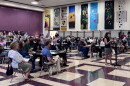 About 150 people in chairs listen to a woman standing at a podium inside an elementary school gymnasium, with musical posters on the wall.