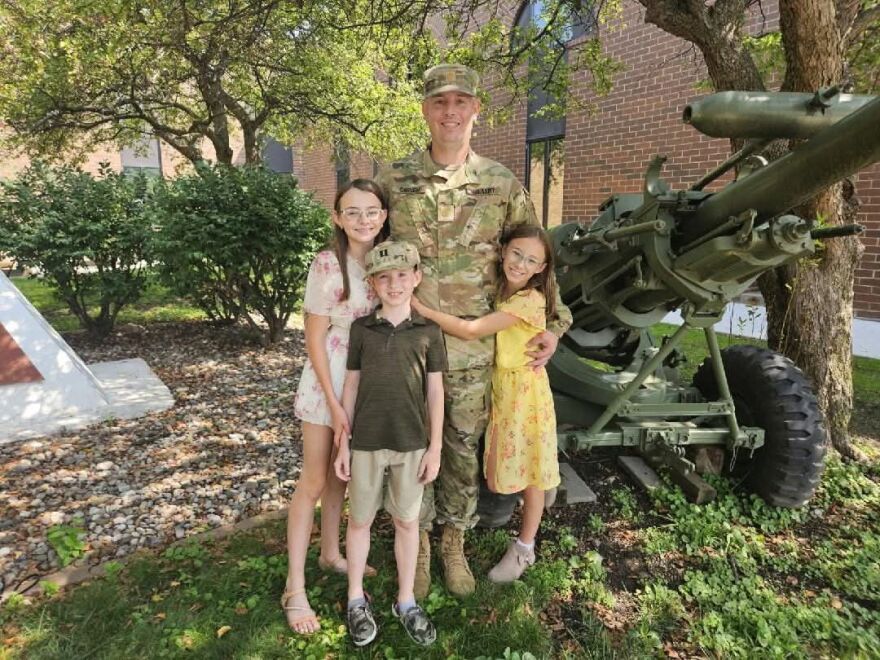 A man in a camouflage military uniform poses for a photo with three young children. They stand near the outdoor display of a military artillery canon.