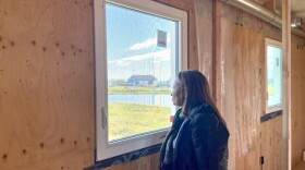 A woman looks out a window from inside a building with plywood on the interior walls.