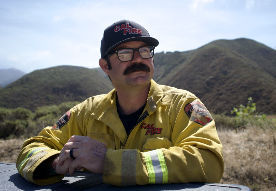 A man in a firefighting jacket with a mustache looks off into the distance with a mountain behind him. 
