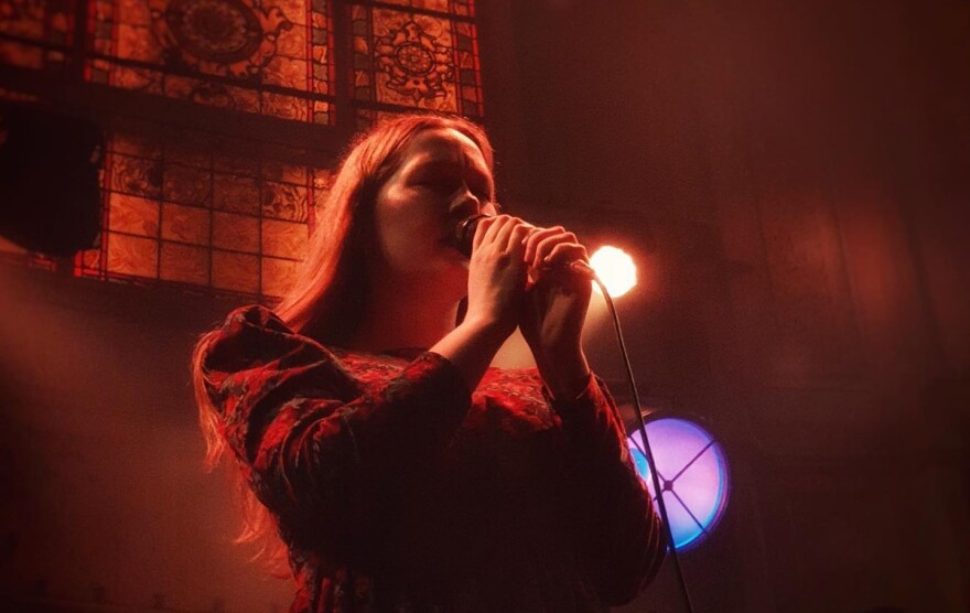  A woman backlit by colored spotlights sings into a microphone while standing in front of a stained-glass window on stage in a darkened music venue.