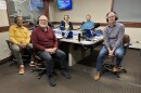 Five smiling people sitting around a table in a radio talk studio: a man front left has short dark hair and a dark mustache and beard and is wearing a yellow shirt, tan pants, white socks, and brown shoes; a man front center is bald and has a white mustache and beard and is wearing a maroon long-sleeved shirt, black pants, black socks, and black shoes; a man back left has short gray hair and is wearing a gray hoodie under a black jacket; a man back left has short brown hair and a brown mustache and beard and is wearing a light blue long-sleeved shirt; a man at right has very short dark hair and is wearing a purple and white plaid shirt, jeans, and brown shoes