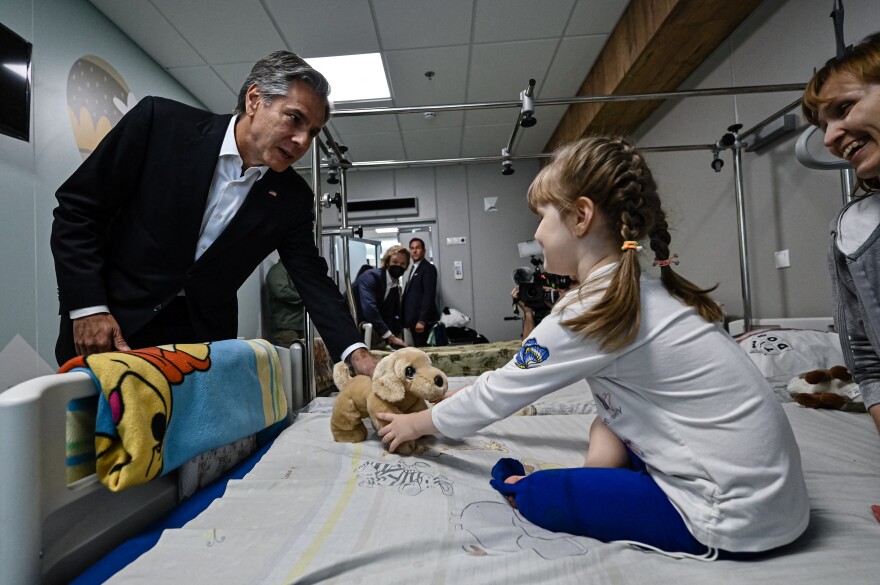 A girl in braids on a hospital bed gets a stuffed doll from U.S. Secretary of State Antony Blinken.