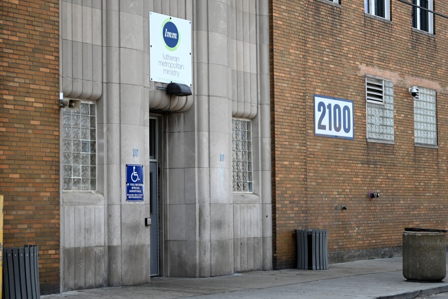 The outside of the Lutheran Metropolitan Ministry has brick walls with stone around the door. The glass block windows are above eyelevel, and a handicapped sign with a button for the door sits just left of the doorway. A large sign with the address, 2100, rests on the side of the building.