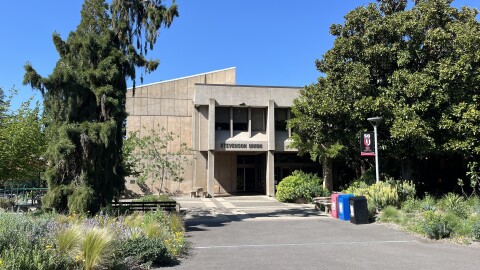 A two-story concrete building, with trees and flowering plants by the entrance. 