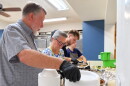 four servers prepare meals in a kitchen. the man closest to the camera with a white beard and hair in a gray short sleeve collared shirt has a stack of plastic plates and bowls. the others behind him are filling the plates and bowls with food
