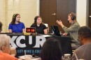 People are seated in an audience watching two women being interviewed by a man at a table. On the table sits an "on air" light and the table is covered by a table cloth that reads KCUR. The camera is positioned behind people in the crowd watching the interview. 