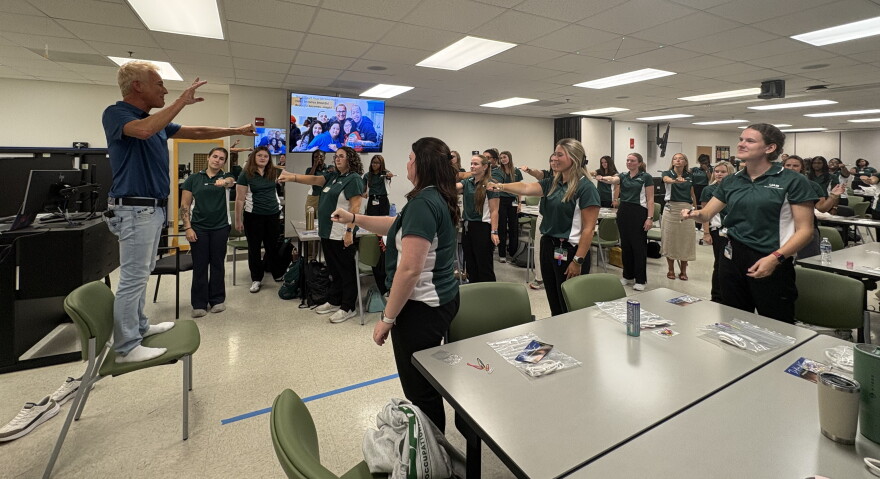 Kevin Spencer stands on a chair with his left arm outstretched to explain a magic trick to a classroom of students at the University of Alabama at Birmingham. The students stand mimicking Spencers position.