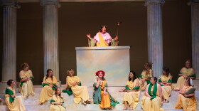 On a stage decorated as a Greek amphitheater, one woman in a Greek costume speaks from a podium to several other women who sit on the steps below. 