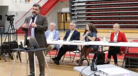 A man in a suit and tie speaks into a microphone in a school gymnasium, with members of the Kentucky Public Service Commission seated at a table behind him.