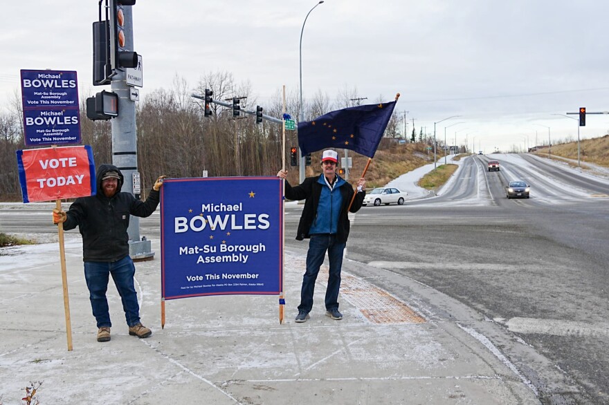 Volunteers hold political campaign signs and an Alaska flag at a large, frosty intersection.