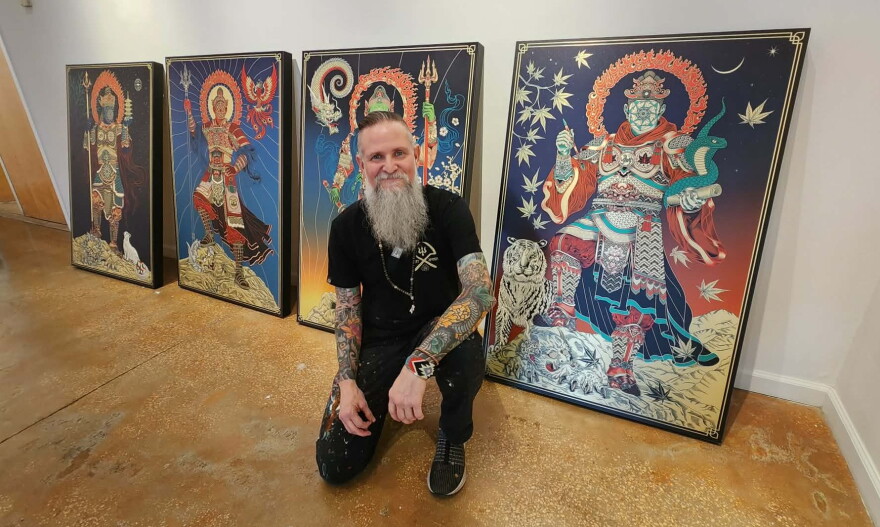 Man with tattoos all over his arms and a long grey beard squats in front of his artwork 